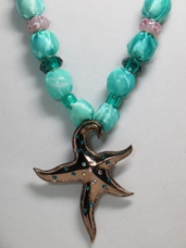 This necklace has a sea foam green silky fabric with a starfish metal pendant of silver tone metal with various green rhinestones embedded in it. The beads are teal green faceted and pony beads and pretty opaque glass beads painted pink and white.