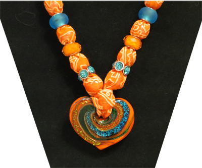 This necklace has a multi-colored heart shaped pendant with orange patterned fabric. The beads are brilliant blue in glass, orange faceted and blue rhinestone. 