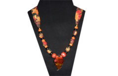 This necklace has a leaf pendant with red and gold glass. The fabric is cotton with leaves of red, gold and orange. The beads are wooden, and black gold tone and orange pony.