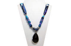 This necklace's pendant is a black faceted teardrop rimed in silver tone metal. The silky fabric is multi-color blue, turquoise, purple and silver. The beads are purple faceted and silver tone metal.