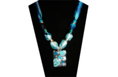 A stunning necklace with variegated turquoise cotton fabric and an oblong pendant with light and dark turquoise raised design on silver tone metal. The beads are silver tone and one with tiny flower and blue rhinestone center.