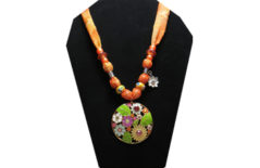 This brightly colored necklace has a round metal medallion covered with yellow, purple, blue, orange, pink and white flowers. The sheer fabric has a pattern of various shades of orange with yellow glass painted beads and orange faceted beads. A cute little white flower with rhinestone center charm compliments the medallion.